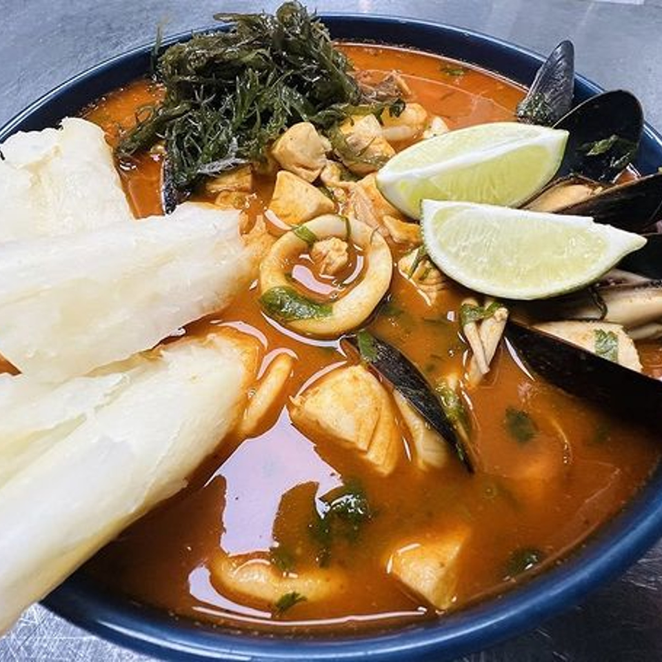 Peruvian seafood soup with mussels, squid, fish, seaweed, yucca, and lime wedges in a blue bowl.