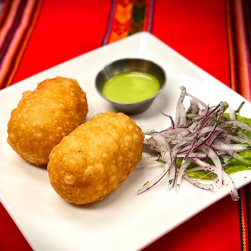 Two golden-brown Peruvian potato croquettes with green sauce and red onion salad on a white plate.