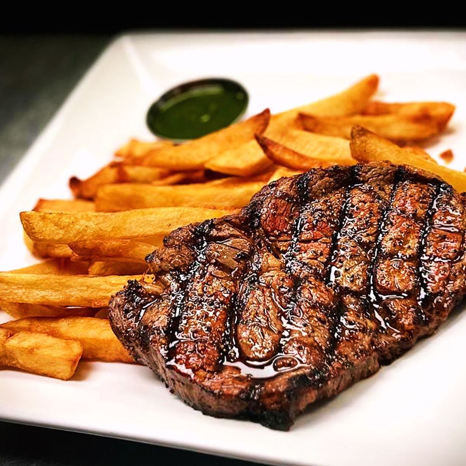 Grilled steak with grill marks, served alongside golden french fries and a small dish of green sauce.